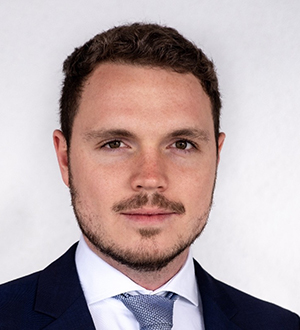 Headshot of a man (Felix Gassert) wearing a black jacket, white collared shirt and a grey tie against a white background