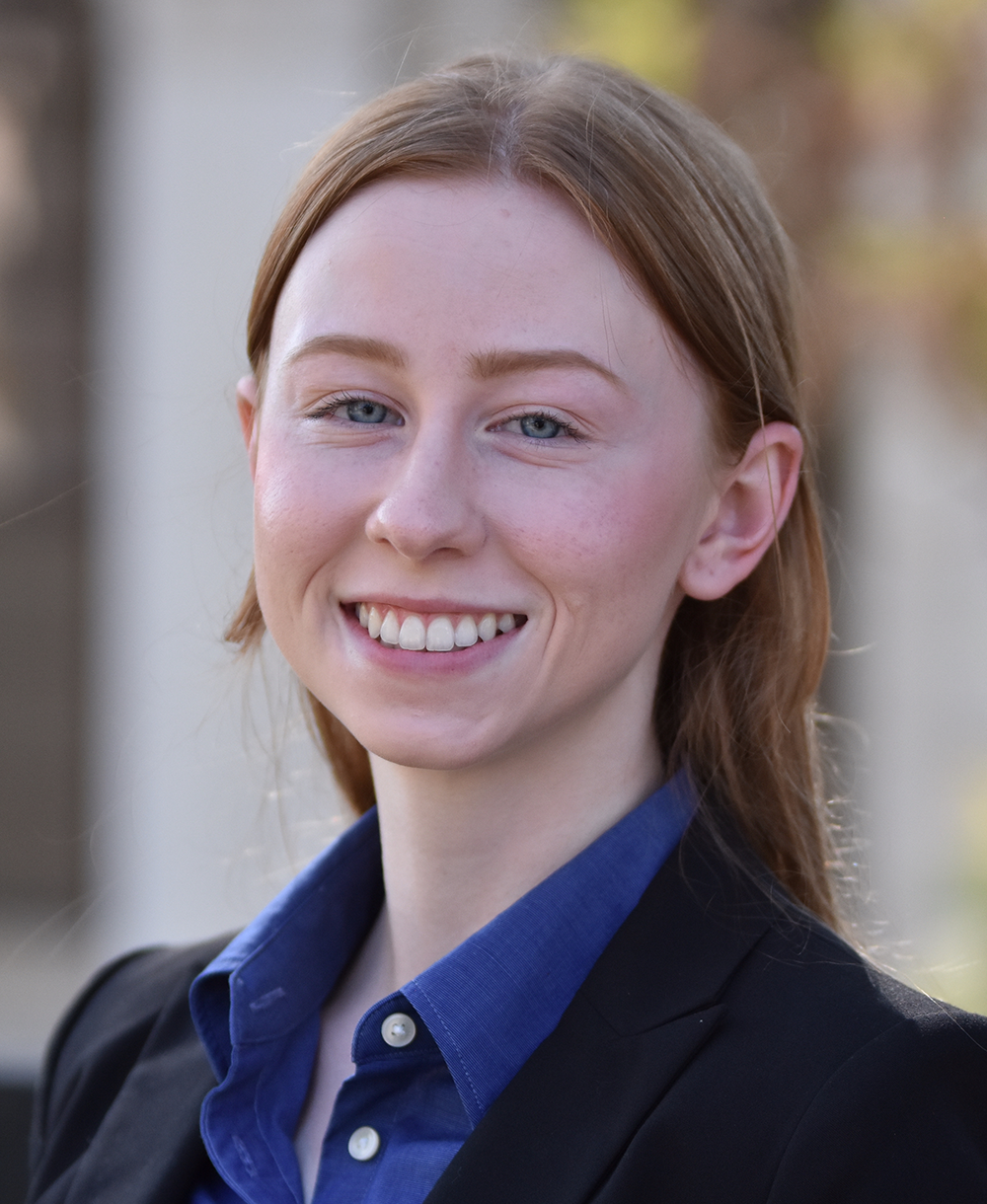 Woman with auburn hair wearing a black jacket and blue collared shirt against an outdoor background