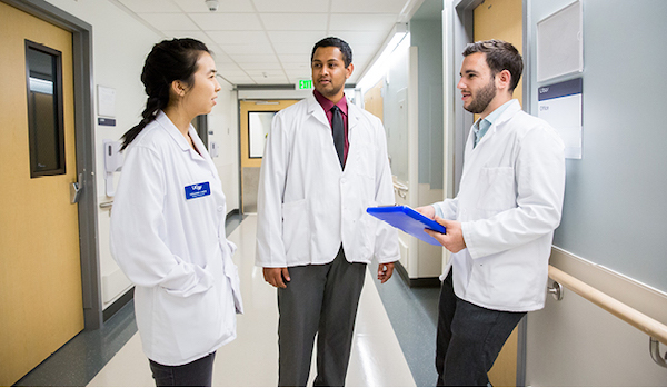 Three students wearing short white coats talk in the hallway at UCSF.