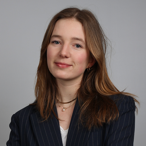 Professional headshot of Emma Duprat with long brown hair, wearing a dark pinstripe blazer against a light gray background.