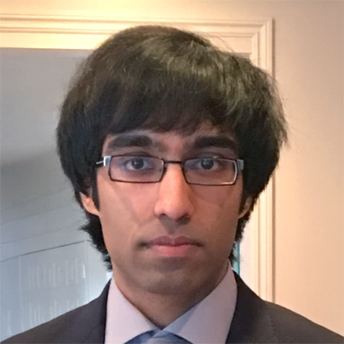 Headshot of a person with dark hair and rectangular glasses, wearing a suit jacket, light-colored dress shirt, and tie, facing the camera against an indoor background.