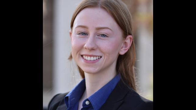 Woman with auburn hair wearing a black jacket and blue collared shirt against an outdoor background