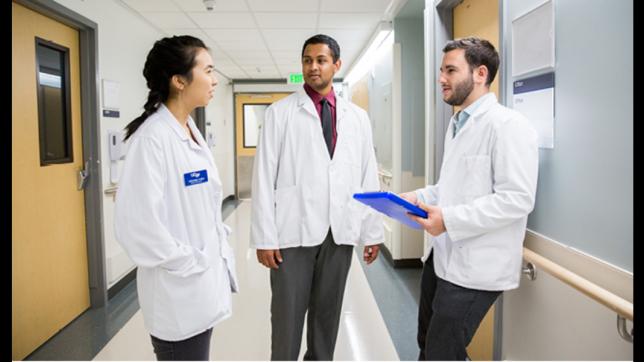Three students wearing short white coats stand talking in the hallway at UCSF.